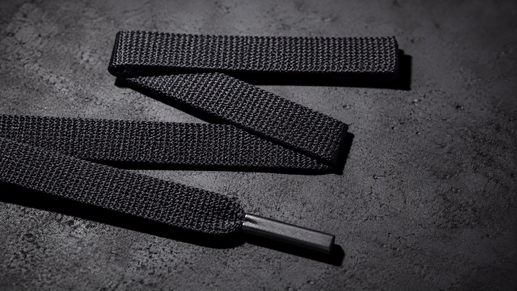 Luxury flat heavy-weave shoelaces by premium brand PrivLace on a raw concrete surface, highlighting custom engraved matte metal tips.